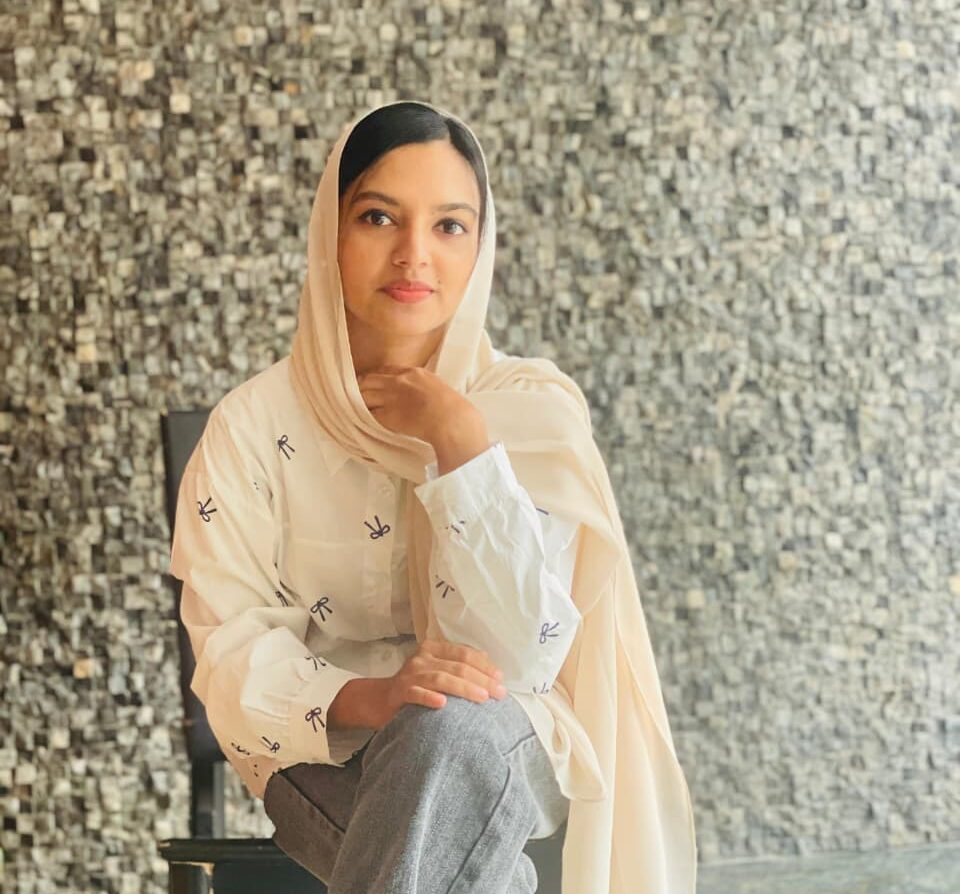 Suhaila V.M, a digital marketing freelancer, sitting in front of a textured gray stone wall, wearing a cream-colored top with a subtle print, gray jeans, and a matching cream-colored hijab.